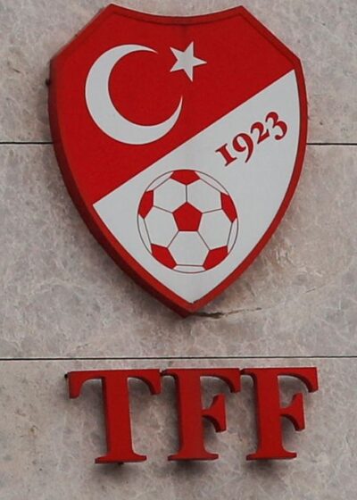 Kuvassa on lähikuva Turkin jalkapalloliiton (TFF) virallisesta kyltistä, joka on kiinnitetty vaaleaan, marmorikuvioituun laattaseinään. Ylimpänä on liiton punainen, kilvenmuotoinen logo. Logossa näkyvät Turkin lipun kuunsirppi ja tähti, perustamisvuosi "1923" sekä perinteinen jalkapallo. Logon alapuolella on näyttävä, punainen kirjainlyhenne "TFF". Tämän alla lukee järjestön täydellinen nimi harmailla kirjaimilla sekä turkiksi ("Türkiye Futbol Federasyonu") että englanniksi ("Turkish Football Federation"), joiden välissä on ohut punainen viiva. Sommitelma on selkeä, virallinen ja ammattimainen.