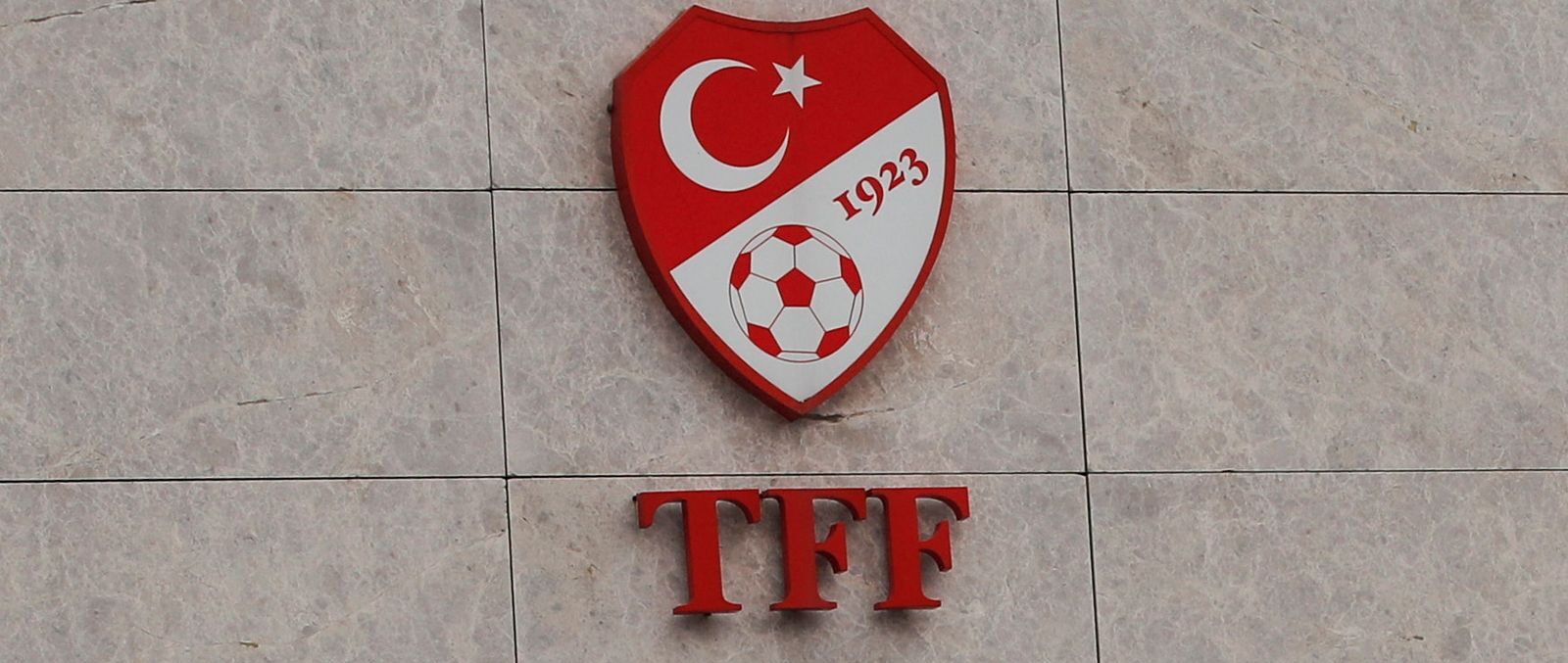 Kuvassa on lähikuva Turkin jalkapalloliiton (TFF) virallisesta kyltistä, joka on kiinnitetty vaaleaan, marmorikuvioituun laattaseinään. Ylimpänä on liiton punainen, kilvenmuotoinen logo. Logossa näkyvät Turkin lipun kuunsirppi ja tähti, perustamisvuosi "1923" sekä perinteinen jalkapallo. Logon alapuolella on näyttävä, punainen kirjainlyhenne "TFF". Tämän alla lukee järjestön täydellinen nimi harmailla kirjaimilla sekä turkiksi ("Türkiye Futbol Federasyonu") että englanniksi ("Turkish Football Federation"), joiden välissä on ohut punainen viiva. Sommitelma on selkeä, virallinen ja ammattimainen.