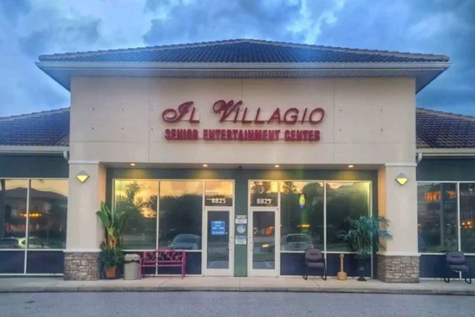 Kuvassa on yksikerroksisen liikerakennuksen julkisivu iltahämärässä. Rakennuksen yläosassa on suuri, punaisena hehkuva kyltti, jossa lukee "Il Villagio" ja sen alla "SENIOR ENTERTAINMENT CENTER", mikä kertoo paikan olevan senioreille suunnattu viihdekeskus. Rakennuksen seinät ovat vaaleat, ja sen alaosassa on kiviverhoilu. Suurista ikkunoista heijastuu pilvinen taivas, ja sisältä loistava lämmin valo luo kutsuvan tunnelman. Sisäänkäynnin edustalla on muutama tuoli, violetti penkki sekä ruukkukasveja. Yleisvaikutelma on rauhallinen ja siisti.
