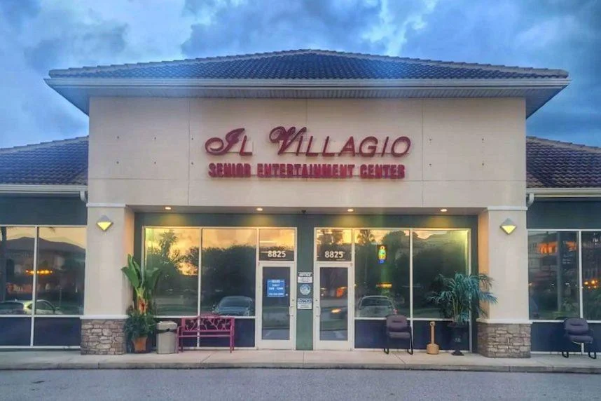 Kuvassa on yksikerroksisen liikerakennuksen julkisivu iltahämärässä. Rakennuksen yläosassa on suuri, punaisena hehkuva kyltti, jossa lukee "Il Villagio" ja sen alla "SENIOR ENTERTAINMENT CENTER", mikä kertoo paikan olevan senioreille suunnattu viihdekeskus. Rakennuksen seinät ovat vaaleat, ja sen alaosassa on kiviverhoilu. Suurista ikkunoista heijastuu pilvinen taivas, ja sisältä loistava lämmin valo luo kutsuvan tunnelman. Sisäänkäynnin edustalla on muutama tuoli, violetti penkki sekä ruukkukasveja. Yleisvaikutelma on rauhallinen ja siisti.