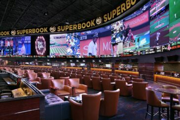 Kuvassa on Westgate Las Vegas -kasinon kuuluisa "SuperBook", yksi maailman suurimmista ja teknologisesti edistyneimmistä urheiluvedonlyöntitiloista. Tilaa hallitsee massiivinen, 4K-videoseinä, joka ulottuu lähes seinästä seinään ja näyttää samanaikaisesti kymmeniä eri live-urheilutapahtumia ja vedonlyöntikertoimia. Hämärästi valaistu katsomo on täynnä satoja mukavia, ruskeita nahkatuoleja ja aitioita, jotka on suunnattu kohti näyttöjä, tarjoten ylellisen ja immersiivisen kokemuksen vedonlyöjille. Tunnelma on dynaaminen ja odottava, vaikka katsomo on juuri nyt melko tyhjä.