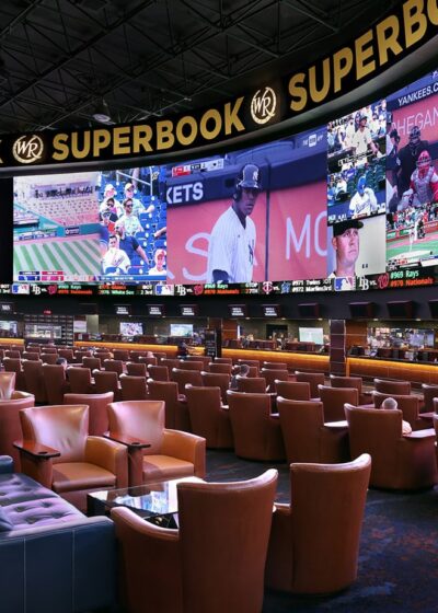 Kuvassa on Westgate Las Vegas -kasinon kuuluisa "SuperBook", yksi maailman suurimmista ja teknologisesti edistyneimmistä urheiluvedonlyöntitiloista. Tilaa hallitsee massiivinen, 4K-videoseinä, joka ulottuu lähes seinästä seinään ja näyttää samanaikaisesti kymmeniä eri live-urheilutapahtumia ja vedonlyöntikertoimia. Hämärästi valaistu katsomo on täynnä satoja mukavia, ruskeita nahkatuoleja ja aitioita, jotka on suunnattu kohti näyttöjä, tarjoten ylellisen ja immersiivisen kokemuksen vedonlyöjille. Tunnelma on dynaaminen ja odottava, vaikka katsomo on juuri nyt melko tyhjä.