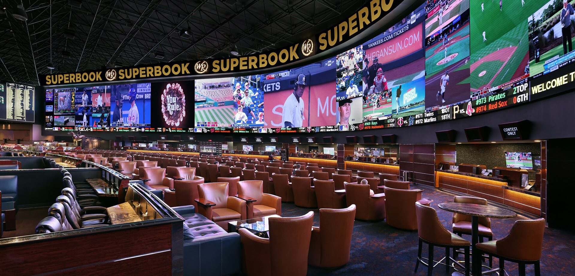 Kuvassa on Westgate Las Vegas -kasinon kuuluisa "SuperBook", yksi maailman suurimmista ja teknologisesti edistyneimmistä urheiluvedonlyöntitiloista. Tilaa hallitsee massiivinen, 4K-videoseinä, joka ulottuu lähes seinästä seinään ja näyttää samanaikaisesti kymmeniä eri live-urheilutapahtumia ja vedonlyöntikertoimia. Hämärästi valaistu katsomo on täynnä satoja mukavia, ruskeita nahkatuoleja ja aitioita, jotka on suunnattu kohti näyttöjä, tarjoten ylellisen ja immersiivisen kokemuksen vedonlyöjille. Tunnelma on dynaaminen ja odottava, vaikka katsomo on juuri nyt melko tyhjä.