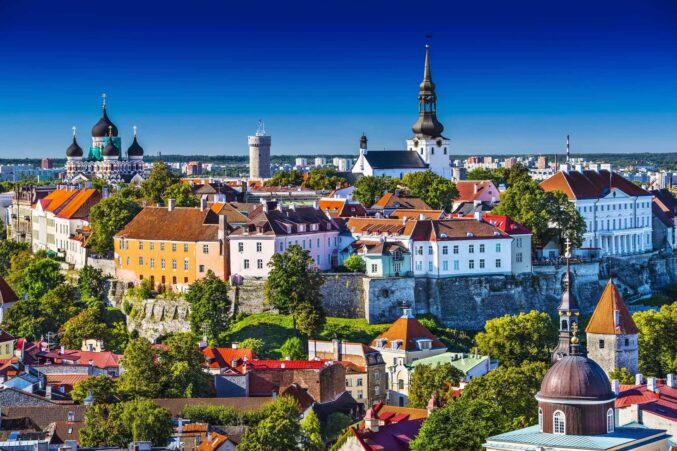 Kuvassa on upea ja aurinkoinen panoraamanäkymä Tallinnan historiallisesta vanhastakaupungista, joka on Unescon maailmanperintökohde. Kuva on otettu kirkkaana päivänä, ja syvänsininen, pilvetön taivas korostaa rakennusten eloisia värejä. Etualalla levittäytyy alempana sijaitsevan vanhankaupungin punaisten tiilikattojen meri, jota täplittävät vehreät puut. Taustalla kohoaa Toompean kukkula, jossa erottuu useita Tallinnan tunnetuimpia maamerkkejä. Vasemmalla on näyttävä, sipulikupolinen Aleksanteri Nevskin katedraali. Keskellä kuvaa ja oikealla näkyvät muun muassa Toomkirik-tuomiokirkon korkea valkoinen torni sekä osa keskiaikaisesta kaupunginmuurista. Näkymä on kuin postikortista, ja se vangitsee täydellisesti Viron pääkaupungin ainutlaatuisen historiallisen tunnelman.