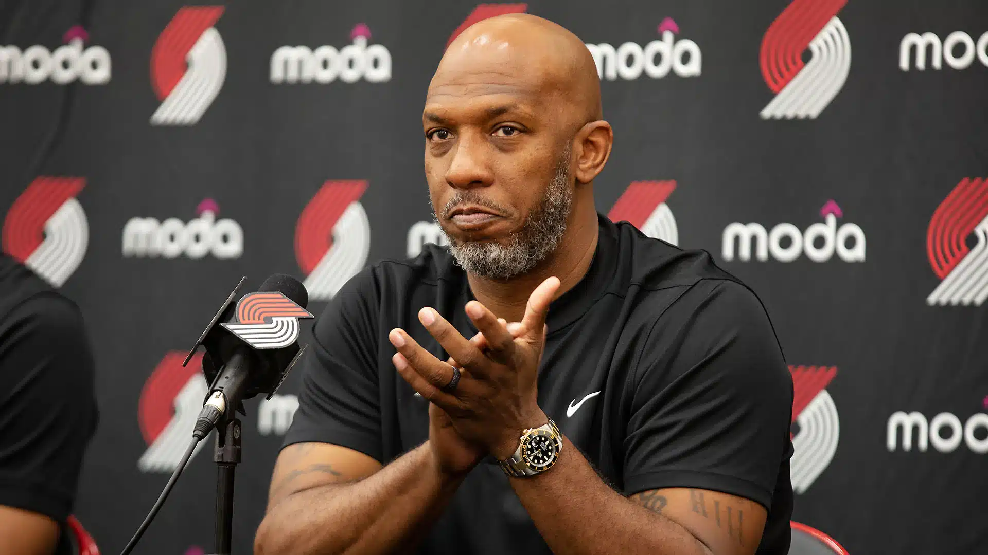 Kuvassa on NBA-koripallojoukkue Portland Trail Blazersin päävalmentaja Chauncey Billups. Hän istuu lehdistötilaisuudessa mikrofonin edessä vakava ja mietteliäs ilme kasvoillaan. Billups on kalju, ja hänellä on harmaantunut parta. Hän on pukeutunut mustaan, Niken logolla varustettuun pikeepaitaan, ja hänen ranteessaan on kultakello. Hänen kätensä ovat koholla ja hän näyttää selittävän tai korostavan jotakin asiaa. Taustalla on musta mainosseinä, joka on täynnä joukkueen tuulimylly-logoa sekä sponsorin "Moda" tekstiä.