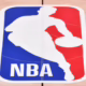 Kuvassa on NBA:n (National Basketball Association) ikoninen logo, joka on maalattu kirkkaalle, vaaleanruskealle koripallokentän parkettilattialle. Logo on pystysuuntainen suorakulmio, jonka vasen puoli on punainen ja oikea sininen. Tämän värijaon päällä on valkoinen siluetti koripalloilijasta kuljettamassa palloa – hahmo perustuu laajalti tunnetusti Jerry Westiin. Logon alaosassa lukee selkeillä, valkoisilla suuraakkosilla "NBA". Kuva on otettu hieman yläviistosta, ja parketin saumat näkyvät logon läpi.