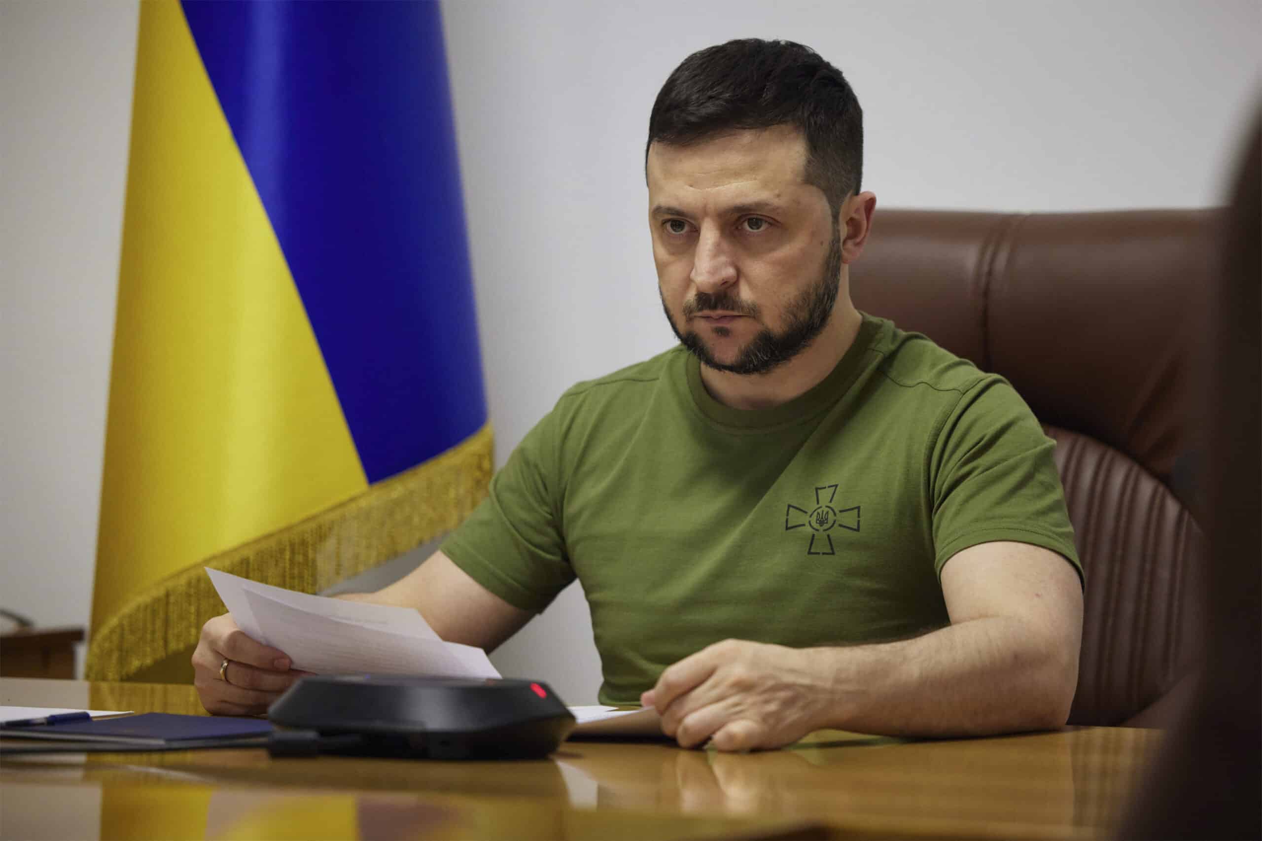 Ukrainan presidentti Zelenskyi istuu puupöydän ääressä edessään papereita ja taustalla Ukrainan lippu