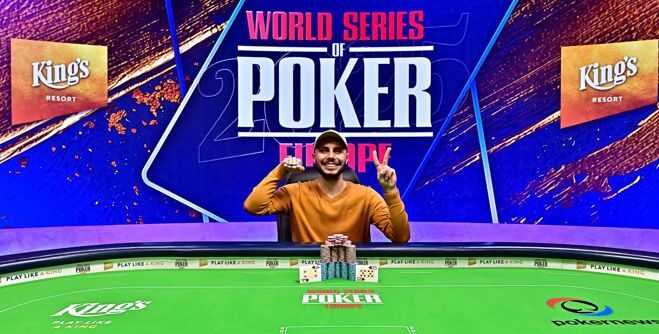 Mies istuu pokeripöydässä päällään keltainen paita ja lippalakki. Mies hymyilee ja esittelee kädessään kultaista ranneketta.