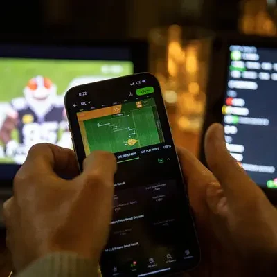 Vedonlyöjä säätää puhelimellaan vedonlyöntisivua ja takana näkyy kaksi tietokoneen näyttöä