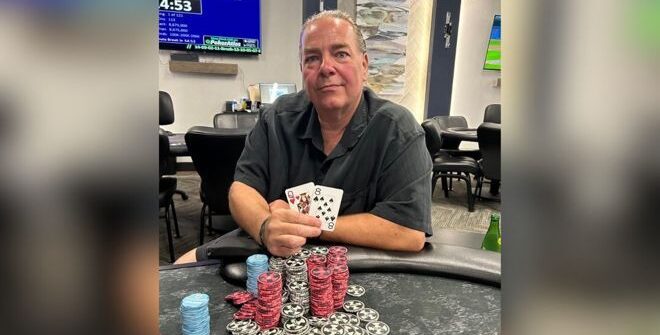 Mies istuu pokeripöydässä. Hänellä on kädessään kaksi pelikorttia. Miehen edessä pöydällä on iso suuri kasa pelimerkkejä.