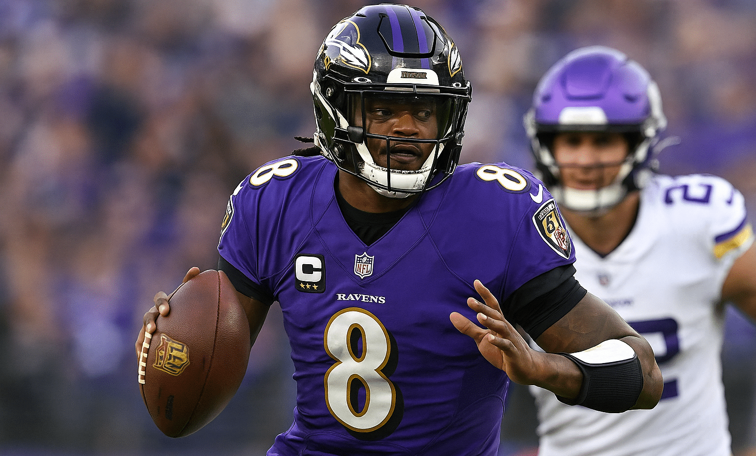 Kaksi miestä pelaavat amerikkalaista jalkapalloa. Toisella on pallo kädessään ja violetti pelipaita. Toinen seisoo takana.