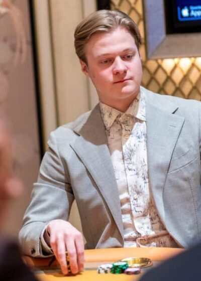 Casimir Seire istuu pokeripöydässä. Hänellä on päällään harmaa puku, jonka alla vaalea kauluspaita. Tausta on kultaista ja ruskeaa.