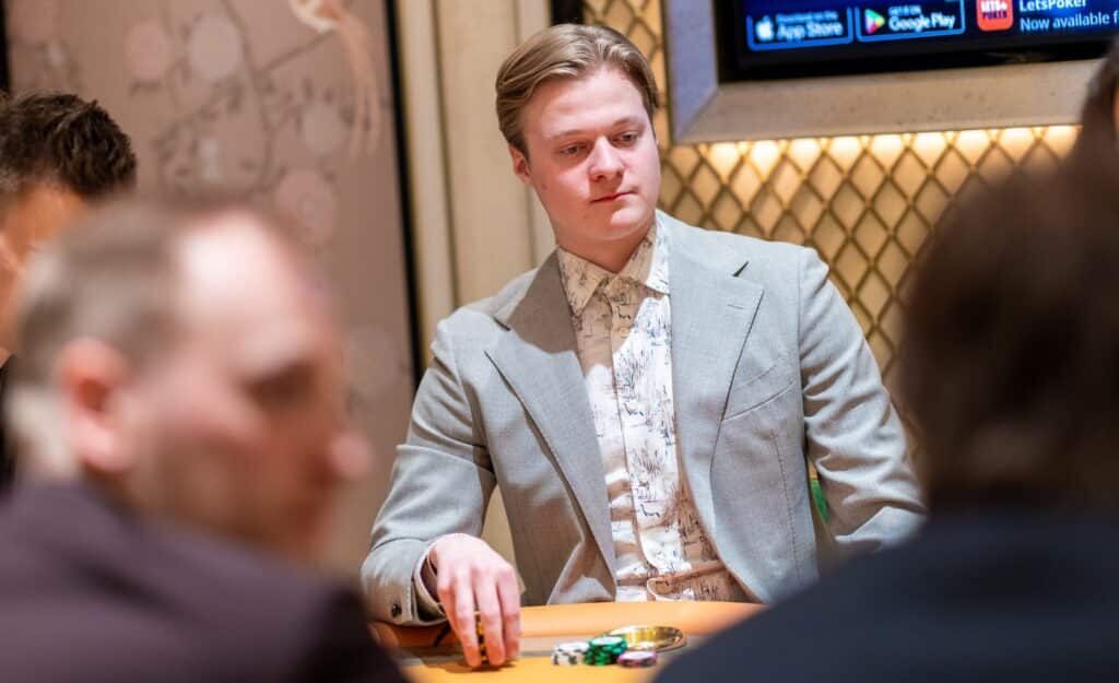 Casimir Seire istuu pokeripöydässä. Hänellä on päällään harmaa puku, jonka alla vaalea kauluspaita. Tausta on kultaista ja ruskeaa.