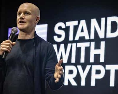 Kuvassa on Coinbasen toimitusjohtaja Brian Armstrong puhumassa lavalla, todennäköisesti kryptovaluuttoihin liittyvässä tapahtumassa. Hän on kalju, pukeutunut tummaan neuleeseen, ja pitää toisessa kädessään mikrofonia samalla kun hän elehtii toisella kädellään korostaakseen asiaansa. Armstrong katsoo yleisöön intensiivisellä ilmeellä. Hänen takanaan on suuri musta näyttö, jossa on suurilla, valkoisilla ja lihavoiduilla kirjaimilla teksti "STAND WITH CRYPTO". Valaistus kohdistuu Armstrongiin, mikä luo kuvaan ammattimaisen ja vaikuttavan tunnelman.