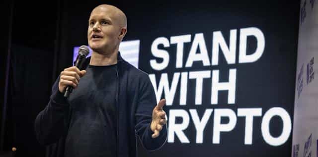 Kuvassa on Coinbasen toimitusjohtaja Brian Armstrong puhumassa lavalla, todennäköisesti kryptovaluuttoihin liittyvässä tapahtumassa. Hän on kalju, pukeutunut tummaan neuleeseen, ja pitää toisessa kädessään mikrofonia samalla kun hän elehtii toisella kädellään korostaakseen asiaansa. Armstrong katsoo yleisöön intensiivisellä ilmeellä. Hänen takanaan on suuri musta näyttö, jossa on suurilla, valkoisilla ja lihavoiduilla kirjaimilla teksti "STAND WITH CRYPTO". Valaistus kohdistuu Armstrongiin, mikä luo kuvaan ammattimaisen ja vaikuttavan tunnelman.