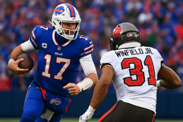 Kaksi miestä pelaavat amerikkalaista jalkapalloa. Toisella on pallo kädessään ja sininen pelipaita. Toinen on selkä edellä edessä.