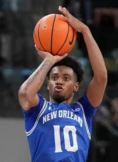 Kuvassa on koripalloilija, joka on juuri heittämässä palloa. Hän käyttää sinistä pelipaitaa, jossa lukee “NEW ORLEANS” ja numero 10. Pelaaja on kohottanut pallon ylös molemmin käsin ja näyttää olevan heittoasennossa, keskittynyt ilme kasvoillaan. Taustalla näkyy hämärtynyt katsomo ja muita pelaajia tai katsojia, mutta tarkennus on selvästi urheilijassa ja itse heittoliikkeessä. Kuva välittää intensiivisen pelitilanteen ja urheilullisen energian.