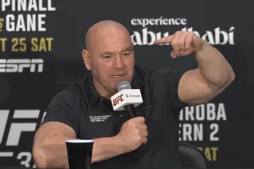 Kuvassa on UFC:n (Ultimate Fighting Championship) toimitusjohtaja Dana White, joka puhuu intensiivisesti lehdistötilaisuudessa. Hän istuu pöydän ääressä, pitää toisessa kädessään UFC-brändättyä mikrofonia ja elehtii voimakkaasti toisella kädellään korostaakseen asiaansa. Hänellä on yllään musta pikeepaita. Whiten takana oleva mainosseinä paljastaa tilaisuuden liittyvän "Aspinall vs. Gane" -otteluun, joka on järjestetty Abu Dhabissa lokakuun 25. päivänä. Whiten ilme on vakava ja latautunut, mikä on hänelle tyypillistä tällaisissa tilaisuuksissa