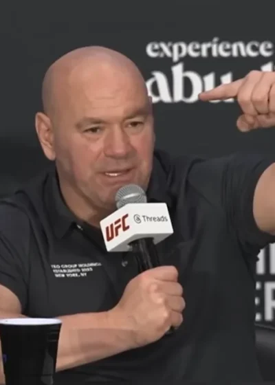 Kuvassa on UFC:n (Ultimate Fighting Championship) toimitusjohtaja Dana White, joka puhuu intensiivisesti lehdistötilaisuudessa. Hän istuu pöydän ääressä, pitää toisessa kädessään UFC-brändättyä mikrofonia ja elehtii voimakkaasti toisella kädellään korostaakseen asiaansa. Hänellä on yllään musta pikeepaita. Whiten takana oleva mainosseinä paljastaa tilaisuuden liittyvän "Aspinall vs. Gane" -otteluun, joka on järjestetty Abu Dhabissa lokakuun 25. päivänä. Whiten ilme on vakava ja latautunut, mikä on hänelle tyypillistä tällaisissa tilaisuuksissa