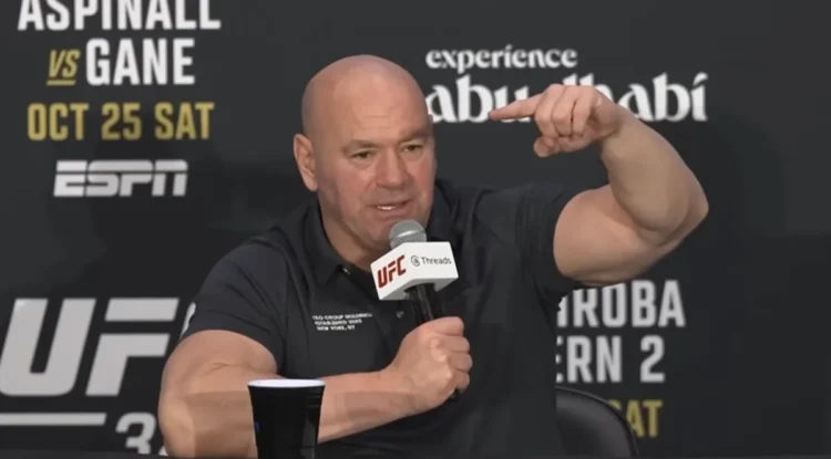 Kuvassa on UFC:n (Ultimate Fighting Championship) toimitusjohtaja Dana White, joka puhuu intensiivisesti lehdistötilaisuudessa. Hän istuu pöydän ääressä, pitää toisessa kädessään UFC-brändättyä mikrofonia ja elehtii voimakkaasti toisella kädellään korostaakseen asiaansa. Hänellä on yllään musta pikeepaita. Whiten takana oleva mainosseinä paljastaa tilaisuuden liittyvän "Aspinall vs. Gane" -otteluun, joka on järjestetty Abu Dhabissa lokakuun 25. päivänä. Whiten ilme on vakava ja latautunut, mikä on hänelle tyypillistä tällaisissa tilaisuuksissa