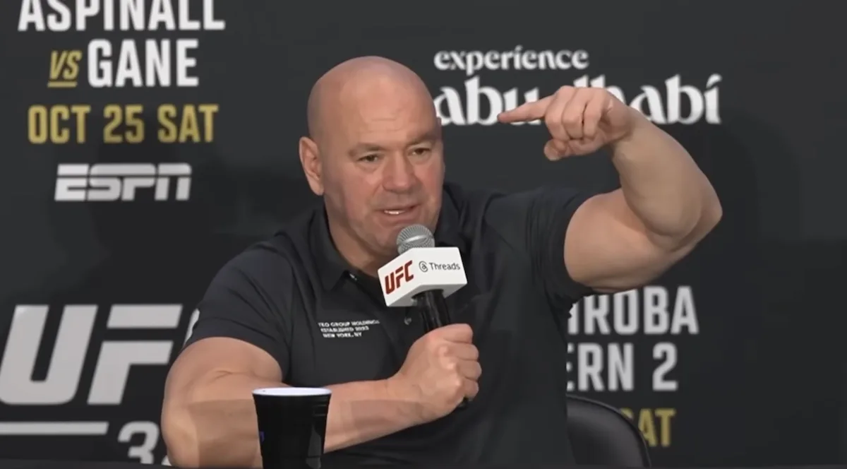 Kuvassa on UFC:n (Ultimate Fighting Championship) toimitusjohtaja Dana White, joka puhuu intensiivisesti lehdistötilaisuudessa. Hän istuu pöydän ääressä, pitää toisessa kädessään UFC-brändättyä mikrofonia ja elehtii voimakkaasti toisella kädellään korostaakseen asiaansa. Hänellä on yllään musta pikeepaita. Whiten takana oleva mainosseinä paljastaa tilaisuuden liittyvän "Aspinall vs. Gane" -otteluun, joka on järjestetty Abu Dhabissa lokakuun 25. päivänä. Whiten ilme on vakava ja latautunut, mikä on hänelle tyypillistä tällaisissa tilaisuuksissa