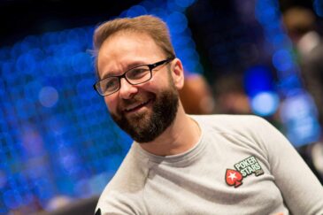 Kuvassa on kanadalainen pokeriammattilainen Daniel Negreanu, yksi maailman tunnetuimmista ja menestyneimmistä pelaajista. Hän hymyilee iloisesti ja rennosti katsellen sivuun. Negreanulla on yllään vaaleanharmaa pitkähihainen paita, jonka rinnassa on hänen pitkäaikaisen sponsorinsa PokerStarsin logo. Hänellä on myös tunnusomaiset silmälasit ja siististi hoidettu parta. Tausta on epätarkka (bokeh-efekti), ja siinä näkyy lukuisia sinisiä valopisteitä, mikä luo kuvaan dynaamisen ja suuren turnaustapahtuman tunnelman. Valokuvan on ottanut Danny Maxwell.