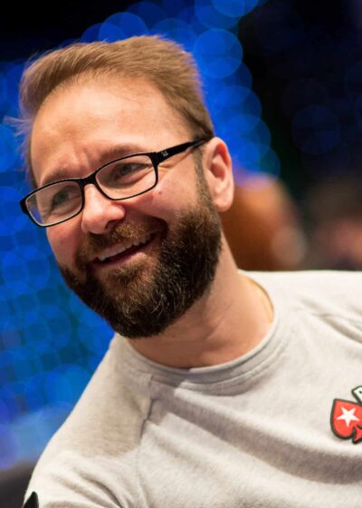 Kuvassa on kanadalainen pokeriammattilainen Daniel Negreanu, yksi maailman tunnetuimmista ja menestyneimmistä pelaajista. Hän hymyilee iloisesti ja rennosti katsellen sivuun. Negreanulla on yllään vaaleanharmaa pitkähihainen paita, jonka rinnassa on hänen pitkäaikaisen sponsorinsa PokerStarsin logo. Hänellä on myös tunnusomaiset silmälasit ja siististi hoidettu parta. Tausta on epätarkka (bokeh-efekti), ja siinä näkyy lukuisia sinisiä valopisteitä, mikä luo kuvaan dynaamisen ja suuren turnaustapahtuman tunnelman. Valokuvan on ottanut Danny Maxwell.