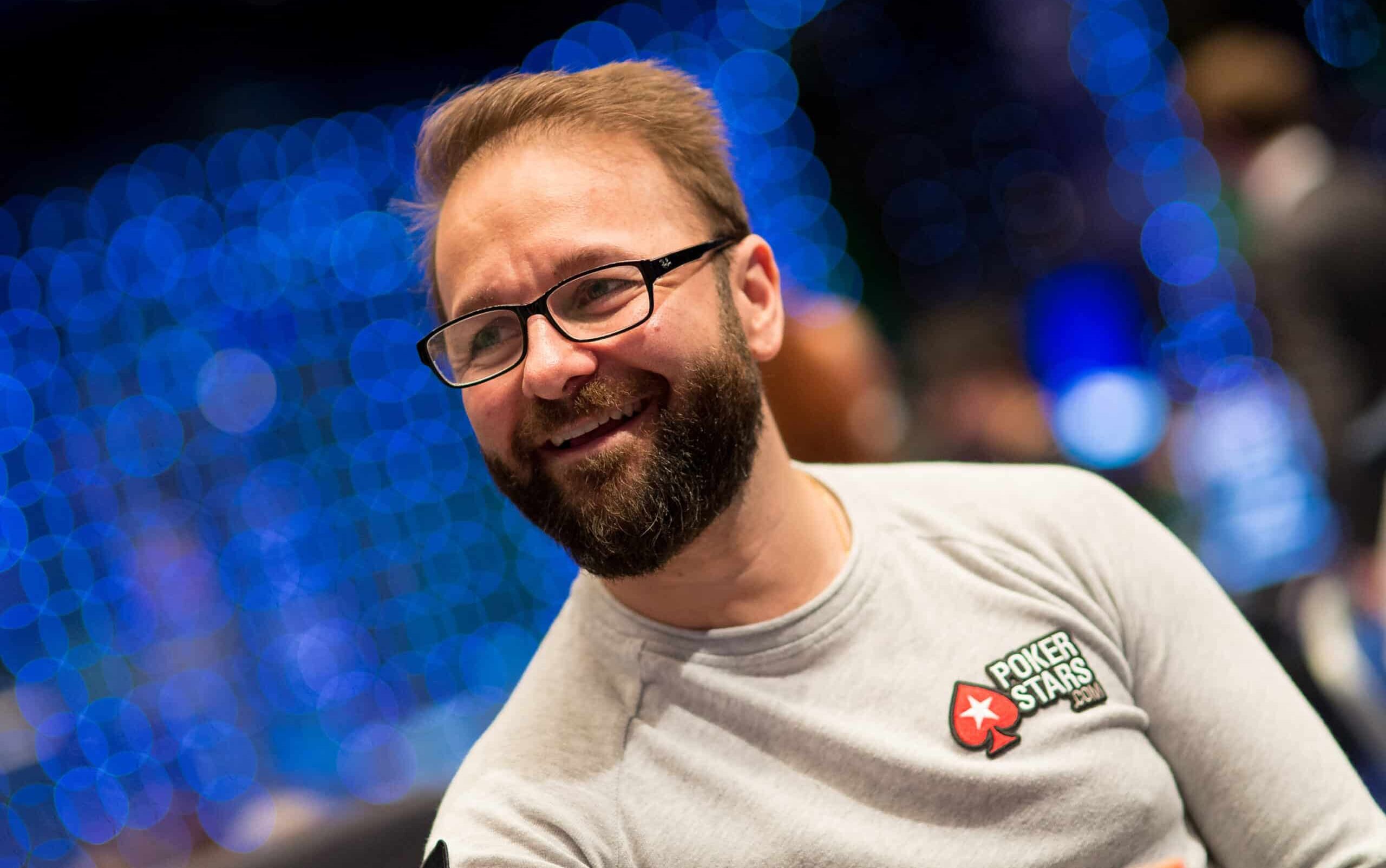 Kuvassa on kanadalainen pokeriammattilainen Daniel Negreanu, yksi maailman tunnetuimmista ja menestyneimmistä pelaajista. Hän hymyilee iloisesti ja rennosti katsellen sivuun. Negreanulla on yllään vaaleanharmaa pitkähihainen paita, jonka rinnassa on hänen pitkäaikaisen sponsorinsa PokerStarsin logo. Hänellä on myös tunnusomaiset silmälasit ja siististi hoidettu parta. Tausta on epätarkka (bokeh-efekti), ja siinä näkyy lukuisia sinisiä valopisteitä, mikä luo kuvaan dynaamisen ja suuren turnaustapahtuman tunnelman. Valokuvan on ottanut Danny Maxwell.