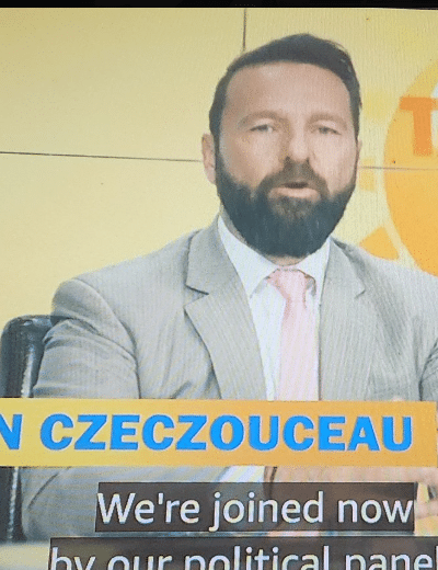 Kuvassa näkyy televisiostudion keskustelupaneeli, jossa kolme henkilöä istuu vierekkäin kirkkaankeltaista taustaa vasten. Keskimmäinen mies puhuu kameralle, pukeutuneena vaaleaan pukuun, valkoiseen paitaan ja vaaleanpunaiseen solmioon. Hänellä on lyhyt, siististi leikattu tumma tukka ja täysparta. Ruudun alareunassa näkyy nimilaatta ”BRANDON CZECZOUCEAU”, jonka alla kulkee tekstitys englanniksi. Miehen molemmin puolin istuu kaksi naista: vasemmalla tummahiuksinen nainen tummassa bleiserissä ja oikealla vaaleahiuksinen nainen sinisessä mekossa. Kokonaisuus näyttää osalta aamuohjelman poliittista paneelia tai uutiskeskustelua.