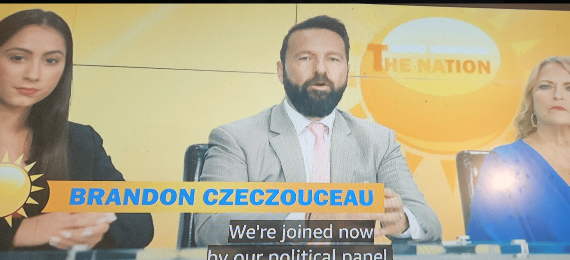 Kuvassa näkyy televisiostudion keskustelupaneeli, jossa kolme henkilöä istuu vierekkäin kirkkaankeltaista taustaa vasten. Keskimmäinen mies puhuu kameralle, pukeutuneena vaaleaan pukuun, valkoiseen paitaan ja vaaleanpunaiseen solmioon. Hänellä on lyhyt, siististi leikattu tumma tukka ja täysparta. Ruudun alareunassa näkyy nimilaatta ”BRANDON CZECZOUCEAU”, jonka alla kulkee tekstitys englanniksi. Miehen molemmin puolin istuu kaksi naista: vasemmalla tummahiuksinen nainen tummassa bleiserissä ja oikealla vaaleahiuksinen nainen sinisessä mekossa. Kokonaisuus näyttää osalta aamuohjelman poliittista paneelia tai uutiskeskustelua.