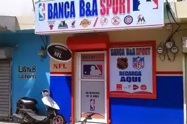 Kuvassa näkyy pienen urheiluvedonlyöntipisteen tai pelibaarin sisäänkäynti, jonka nimi on BANCA B&A SPORT. Julkisivu on maalattu sinisen, valkoisen ja punaisen sävyillä, ja sen yläpuolella on suuri kyltti, jossa näkyy useiden yhdysvaltalaisten urheiluliigojen logot, kuten NBA, MLB ja muiden joukkueiden tunnuksia. Oven vieressä on mainoksia NFL:stä, NHL:stä ja muista urheilusarjoista, ja teksti “RECARGA AQUI” viittaa puhelin- tai palvelulatauksiin, joita voi tehdä samassa paikassa. Oven alareunassa lukee espanjaksi, ettei alaikäisille ole pääsyä. Rakennuksen edessä on mopo pysäköitynä ja vasemmalla puolella näkyy siniseksi maalattu seinä, jossa on teksti “LISTA B LAS FLORES”. Kokonaisuus antaa vaikutelman Karibian tai Latinalaisen Amerikan katunäkymästä.