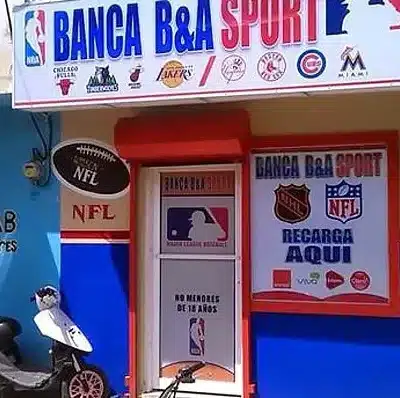 Kuvassa näkyy pienen urheiluvedonlyöntipisteen tai pelibaarin sisäänkäynti, jonka nimi on BANCA B&A SPORT. Julkisivu on maalattu sinisen, valkoisen ja punaisen sävyillä, ja sen yläpuolella on suuri kyltti, jossa näkyy useiden yhdysvaltalaisten urheiluliigojen logot, kuten NBA, MLB ja muiden joukkueiden tunnuksia. Oven vieressä on mainoksia NFL:stä, NHL:stä ja muista urheilusarjoista, ja teksti “RECARGA AQUI” viittaa puhelin- tai palvelulatauksiin, joita voi tehdä samassa paikassa. Oven alareunassa lukee espanjaksi, ettei alaikäisille ole pääsyä. Rakennuksen edessä on mopo pysäköitynä ja vasemmalla puolella näkyy siniseksi maalattu seinä, jossa on teksti “LISTA B LAS FLORES”. Kokonaisuus antaa vaikutelman Karibian tai Latinalaisen Amerikan katunäkymästä.