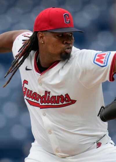 Kuvassa on Cleveland Guardians -joukkueen baseball-syöttäjä intensiivisessä heittoliikkeessä kesken ottelun. Pelaajalla on yllään joukkueen valkoinen kotipeliasu, jonka rinnuksessa lukee punaisella kaunokirjoituksella "Guardians". Päässään hänellä on punainen lippalakki, jossa on C-kirjain, ja lakin alta valuvat pitkät rastahiukset. Hänen oikea kätensä on virittynyt kauas taakse pitelemään palloa, kun taas vasemmassa kädessä oleva musta räpylä osoittaa eteenpäin kohti (kuvan ulkopuolella olevaa) lyöjää. Pelaajan ilme on äärimmäisen keskittynyt ja dynaaminen. Tausta koostuu epätarkoista sinisistä stadionin istuimista, mikä korostaa urheilusuorituksen vauhtia.