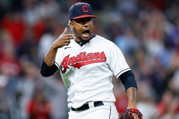 Kuvassa on baseball-pelaaja Cleveland Indians -joukkueen (nykyinen Guardians) peliasussa. Hän on vangittu erittäin intensiiviseen ja tunteikkaaseen hetkeen kesken pelin. Pelaajalla on yllään valkoinen pelipaita, jossa on punainen 'Indians'-teksti, sekä laivastonsininen C-kirjaimella varustettu lippalakki. Hänen ilmeensä on latautunut: hän huutaa suu auki ja osoittaa samalla sormellaan päätään tai korvaansa, mikä on voimakas tuuletus tai reaktio yleisön pauhuun. Taustalla näkyy sumennettuna täysi stadionkatsomo, mikä korostaa pelaajan olevan hetken keskipisteessä. Kuva on täynnä adrenaliinia ja urheilun intohimoa.