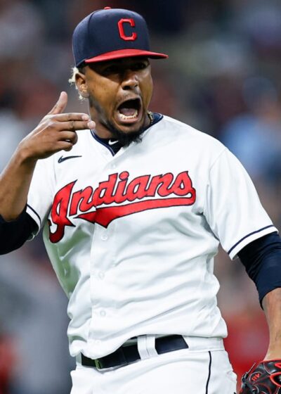 Kuvassa on baseball-pelaaja Cleveland Indians -joukkueen (nykyinen Guardians) peliasussa. Hän on vangittu erittäin intensiiviseen ja tunteikkaaseen hetkeen kesken pelin. Pelaajalla on yllään valkoinen pelipaita, jossa on punainen 'Indians'-teksti, sekä laivastonsininen C-kirjaimella varustettu lippalakki. Hänen ilmeensä on latautunut: hän huutaa suu auki ja osoittaa samalla sormellaan päätään tai korvaansa, mikä on voimakas tuuletus tai reaktio yleisön pauhuun. Taustalla näkyy sumennettuna täysi stadionkatsomo, mikä korostaa pelaajan olevan hetken keskipisteessä. Kuva on täynnä adrenaliinia ja urheilun intohimoa.