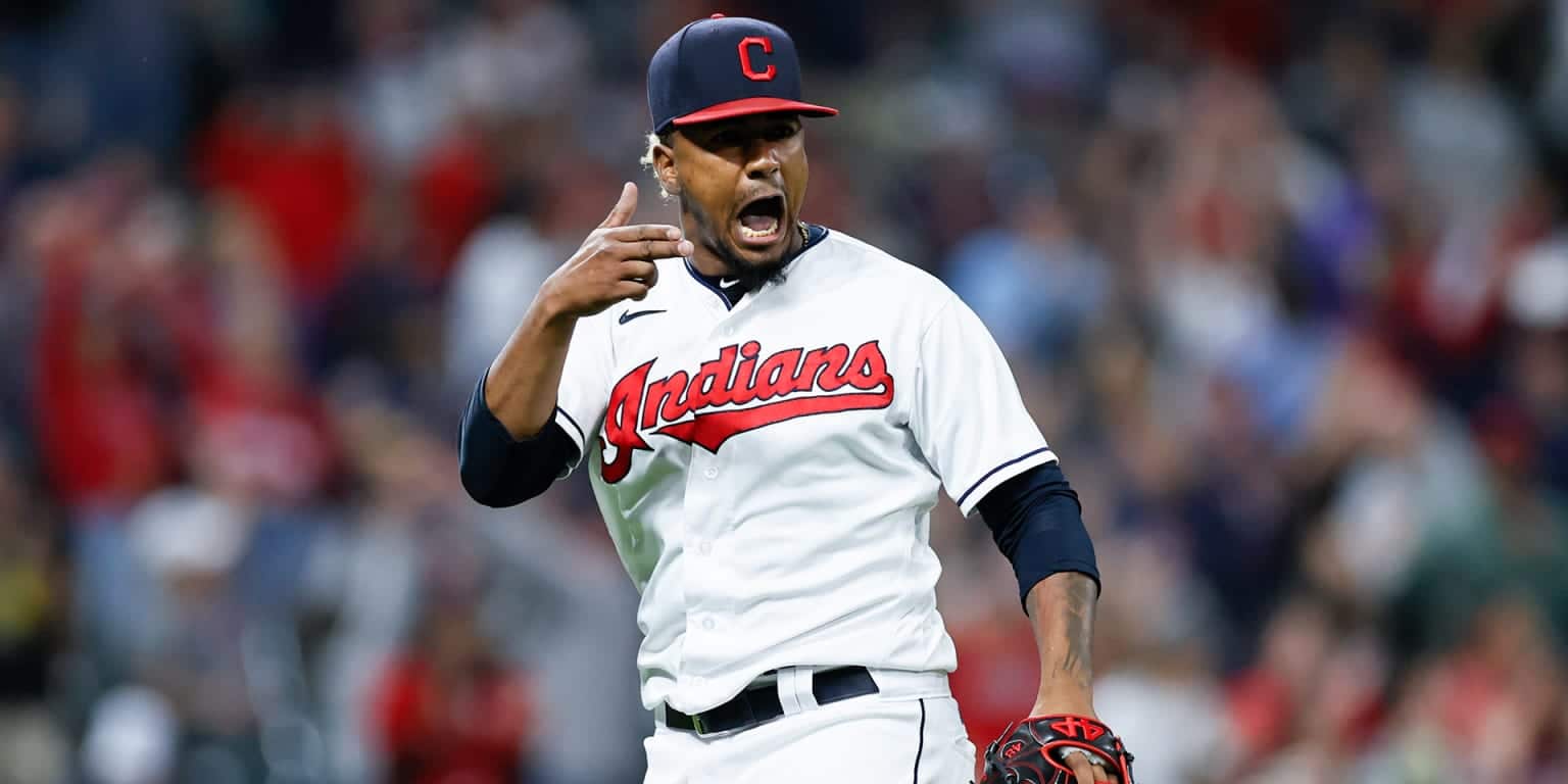 Kuvassa on baseball-pelaaja Cleveland Indians -joukkueen (nykyinen Guardians) peliasussa. Hän on vangittu erittäin intensiiviseen ja tunteikkaaseen hetkeen kesken pelin. Pelaajalla on yllään valkoinen pelipaita, jossa on punainen 'Indians'-teksti, sekä laivastonsininen C-kirjaimella varustettu lippalakki. Hänen ilmeensä on latautunut: hän huutaa suu auki ja osoittaa samalla sormellaan päätään tai korvaansa, mikä on voimakas tuuletus tai reaktio yleisön pauhuun. Taustalla näkyy sumennettuna täysi stadionkatsomo, mikä korostaa pelaajan olevan hetken keskipisteessä. Kuva on täynnä adrenaliinia ja urheilun intohimoa.