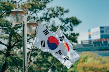 Kuvassa on kaksi Etelä-Korean lippua, jotka on kiinnitetty metalliseen, kaksihaaraiseen katuvalopylvääseen. Liput riippuvat viistosti alaspäin. Etualalla ja lippujen takana näkyy vihreitä havupuita. Taustalla siintää epätarkkana kaupunkimaisema, jossa erottuu vaaleita rakennuksia ja mainoskylttejä. Sää on aurinkoinen ja taivas on kirkkaansininen, mikä luo kuvaan raikkaan päivänvalon tunnelman.