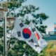 Kuvassa on kaksi Etelä-Korean lippua, jotka on kiinnitetty metalliseen, kaksihaaraiseen katuvalopylvääseen. Liput riippuvat viistosti alaspäin. Etualalla ja lippujen takana näkyy vihreitä havupuita. Taustalla siintää epätarkkana kaupunkimaisema, jossa erottuu vaaleita rakennuksia ja mainoskylttejä. Sää on aurinkoinen ja taivas on kirkkaansininen, mikä luo kuvaan raikkaan päivänvalon tunnelman.