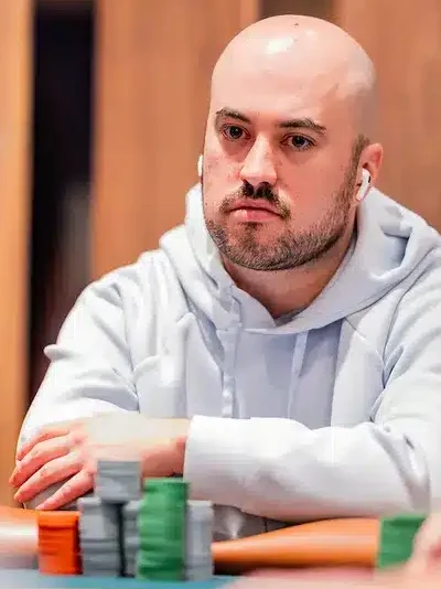 Kuvassa on kalju, parrakas mies, joka istuu pelipöydän ääressä äärimmäisen keskittyneenä. Hänellä on yllään vaalea huppari ja korvissaan valkoiset, langattomat nappikuulokkeet. Miehen ilme on vakava ja tuima, ja hän nojaa eteenpäin kädet ristissä pöydällä. Hänen edessään sinivihreällä pöydällä on pieniä pinoja harmaita, vihreitä ja oransseja pelimerkkejä. Tausta on sumea, mutta siellä näkyy muita ihmisiä, mikä viittaa vahvasti pokeriturnaukseen. Kuva vangitsee täydellisesti hetken, joka on täynnä intensiivistä pohdintaa ja keskittymistä.