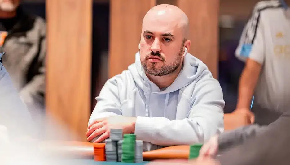 Kuvassa on kalju, parrakas mies, joka istuu pelipöydän ääressä äärimmäisen keskittyneenä. Hänellä on yllään vaalea huppari ja korvissaan valkoiset, langattomat nappikuulokkeet. Miehen ilme on vakava ja tuima, ja hän nojaa eteenpäin kädet ristissä pöydällä. Hänen edessään sinivihreällä pöydällä on pieniä pinoja harmaita, vihreitä ja oransseja pelimerkkejä. Tausta on sumea, mutta siellä näkyy muita ihmisiä, mikä viittaa vahvasti pokeriturnaukseen. Kuva vangitsee täydellisesti hetken, joka on täynnä intensiivistä pohdintaa ja keskittymistä.