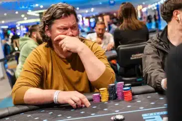 Kuvassa on pokerinpelaaja syvissä mietteissään kesken jännittävän hetken "888LIVE Barcelona" -turnauksessa. Keski-ikäinen, tummahiuksinen mies istuu pelipöydän ääressä ja näyttää erittäin keskittyneeltä tai jopa huolestuneelta. Hän pitää toista kättään suunsa edessä, ja hänen katseensa on suunnattu mietteliäänä sivuun. Hänen edessään mustalla pöydällä on useita pinoja keltaisia, purppuranpunaisia ja mustia pelimerkkejä. Tausta on kirkkaasti valaistu kasinosali täynnä muita pelaajia. Kuva vangitsee erinomaisesti sen intensiivisen paineen ja psykologisen pelin, joka liittyy korkean panoksen pokeriin.