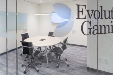 Kuvassa on Evolution Gaming -yhtiön moderni ja siisti neuvotteluhuone, joka nähdään suuren lasiseinän läpi. Lasiseinään on teipattu yrityksen logo sekä nimi "Evolution Gaming" suurilla, tummilla kirjaimilla. Huone on kalustettu minimalistisesti: sen keskellä on pyöreä, vaalea pöytä, jota ympäröi viisi mustaa, pyörillä varustettua toimistotuolia. Takaseinällä on seinään kiinnitetty televisioruutu ja vasemmalla seinustalla kaksi valkotaulua. Lattia on harmaata puukuvioitua laminaattia, ja katosta roikkuu moderni rengasvalaisin. Yleisilme on erittäin ammattimainen ja puhdas.