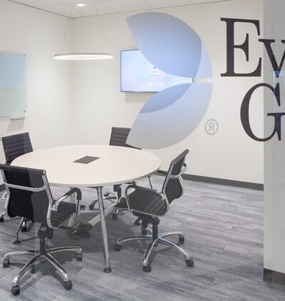 Kuvassa on Evolution Gaming -yhtiön moderni ja siisti neuvotteluhuone, joka nähdään suuren lasiseinän läpi. Lasiseinään on teipattu yrityksen logo sekä nimi "Evolution Gaming" suurilla, tummilla kirjaimilla. Huone on kalustettu minimalistisesti: sen keskellä on pyöreä, vaalea pöytä, jota ympäröi viisi mustaa, pyörillä varustettua toimistotuolia. Takaseinällä on seinään kiinnitetty televisioruutu ja vasemmalla seinustalla kaksi valkotaulua. Lattia on harmaata puukuvioitua laminaattia, ja katosta roikkuu moderni rengasvalaisin. Yleisilme on erittäin ammattimainen ja puhdas.