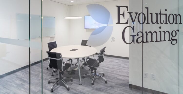 Kuvassa on Evolution Gaming -yhtiön moderni ja siisti neuvotteluhuone, joka nähdään suuren lasiseinän läpi. Lasiseinään on teipattu yrityksen logo sekä nimi "Evolution Gaming" suurilla, tummilla kirjaimilla. Huone on kalustettu minimalistisesti: sen keskellä on pyöreä, vaalea pöytä, jota ympäröi viisi mustaa, pyörillä varustettua toimistotuolia. Takaseinällä on seinään kiinnitetty televisioruutu ja vasemmalla seinustalla kaksi valkotaulua. Lattia on harmaata puukuvioitua laminaattia, ja katosta roikkuu moderni rengasvalaisin. Yleisilme on erittäin ammattimainen ja puhdas.