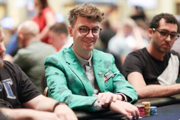 Kuvassa on tunnettu irlantilainen pokerinpelaaja ja striimaaja Fintan "easywithaces" Hand, joka hymyilee lämpimästi kameralle kesken turnauspelin. Hänen olemuksensa on erittäin persoonallinen: hänellä on yllään kirkkaanvihreä puvuntakki, joka on kauttaaltaan koristeltu tummemmilla neliapilakuvioilla, viitaten hänen irlantilaiseen perintöönsä. Takin rinnassa on PokerStars-logo. Hänellä on myös pyöreät silmälasit ja kiharat hiukset. Hand istuu pelipöydän ääressä, kädet pienen pelimerkkipinon edessä. Tausta on sumea kasinosali. Kuvan on ottanut valokuvaaja Carlos Monti.