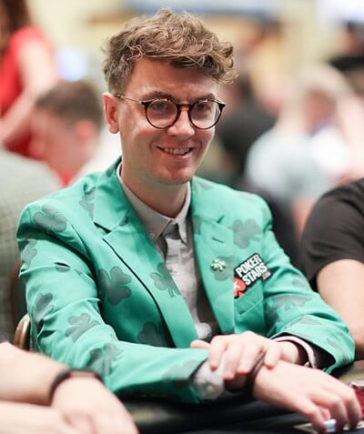 Kuvassa on tunnettu irlantilainen pokerinpelaaja ja striimaaja Fintan "easywithaces" Hand, joka hymyilee lämpimästi kameralle kesken turnauspelin. Hänen olemuksensa on erittäin persoonallinen: hänellä on yllään kirkkaanvihreä puvuntakki, joka on kauttaaltaan koristeltu tummemmilla neliapilakuvioilla, viitaten hänen irlantilaiseen perintöönsä. Takin rinnassa on PokerStars-logo. Hänellä on myös pyöreät silmälasit ja kiharat hiukset. Hand istuu pelipöydän ääressä, kädet pienen pelimerkkipinon edessä. Tausta on sumea kasinosali. Kuvan on ottanut valokuvaaja Carlos Monti.