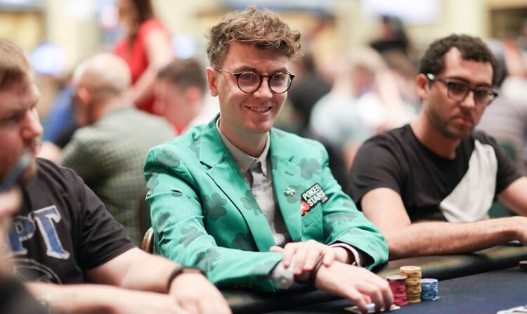 Kuvassa on tunnettu irlantilainen pokerinpelaaja ja striimaaja Fintan "easywithaces" Hand, joka hymyilee lämpimästi kameralle kesken turnauspelin. Hänen olemuksensa on erittäin persoonallinen: hänellä on yllään kirkkaanvihreä puvuntakki, joka on kauttaaltaan koristeltu tummemmilla neliapilakuvioilla, viitaten hänen irlantilaiseen perintöönsä. Takin rinnassa on PokerStars-logo. Hänellä on myös pyöreät silmälasit ja kiharat hiukset. Hand istuu pelipöydän ääressä, kädet pienen pelimerkkipinon edessä. Tausta on sumea kasinosali. Kuvan on ottanut valokuvaaja Carlos Monti.