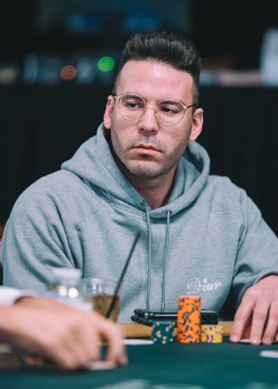 Kuvassa on pokeripöydän ääressä istuva henkilö, joka keskittyy tilanteeseen vakavan ja tarkkaavaisen näköisenä. Hänellä on päällään vaaleanharmaa huppari ja pyöreät silmälasit, jotka korostavat hänen ilmettään. Edessä pöydällä on useita värikkäitä pelimerkkejä pinossa sekä juomapullo ja muutamia muita pieniä esineitä. Tausta on hämärä ja pehmeästi epätarkka, mikä tuo huomion pelaajaan ja pelitilanteeseen. Vasemmalla näkyy toinen pelaaja osittain, mutta kuva keskittyy selvästi keskellä istuvaan henkilöön ja tämän ilmeeseen, joka heijastaa turnaustilanteen vakavuutta.