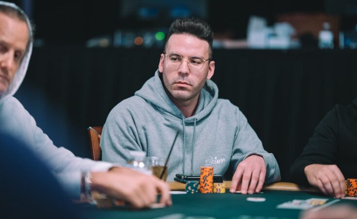 Kuvassa on pokeripöydän ääressä istuva henkilö, joka keskittyy tilanteeseen vakavan ja tarkkaavaisen näköisenä. Hänellä on päällään vaaleanharmaa huppari ja pyöreät silmälasit, jotka korostavat hänen ilmettään. Edessä pöydällä on useita värikkäitä pelimerkkejä pinossa sekä juomapullo ja muutamia muita pieniä esineitä. Tausta on hämärä ja pehmeästi epätarkka, mikä tuo huomion pelaajaan ja pelitilanteeseen. Vasemmalla näkyy toinen pelaaja osittain, mutta kuva keskittyy selvästi keskellä istuvaan henkilöön ja tämän ilmeeseen, joka heijastaa turnaustilanteen vakavuutta.