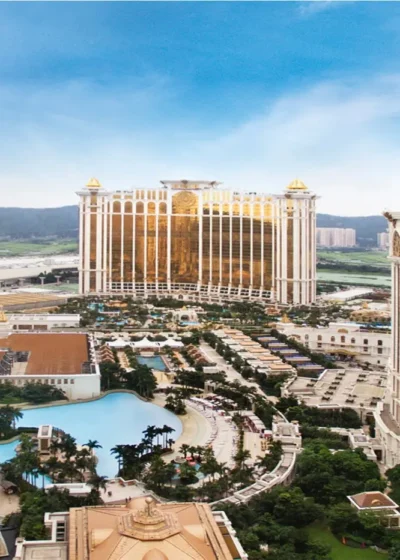 Kuvassa on henkeäsalpaava panoraamanäkymä valtavasta ja äärimmäisen ylellisestä lomakeskuksesta, joka tunnetaan nimellä Galaxy Macau. Kuvaa reunustavat etualalla kaksi massiivista, palatsimaista hotellitornia. Niiden vaaleaa julkisivua korostavat runsaat kultaiset yksityiskohdat, ja tornien huipulla kiiltävät näyttävät kultaiset kupolit. Tornien väliin jää laaja, trooppinen virkistysalue, jossa erottuu suuria uima-altaita, vehreitä puutarhoja ja palmuryhmiä. Taustalla kohoaa kolmas, kullanvärinen ja jättiläismäinen hotellirakennus. Näkymä huokuu "Aasian Las Vegasin" yltäkylläisyyttä ja luksusta.
